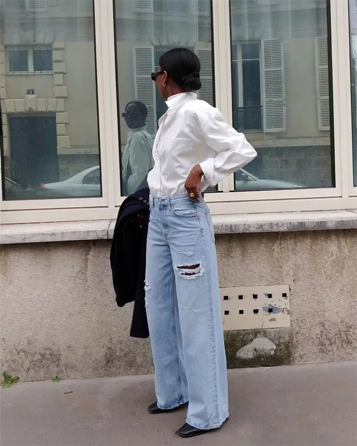 Девушка в потертых синих джинсах, белой рубашке и черных туфлях на каблуках.