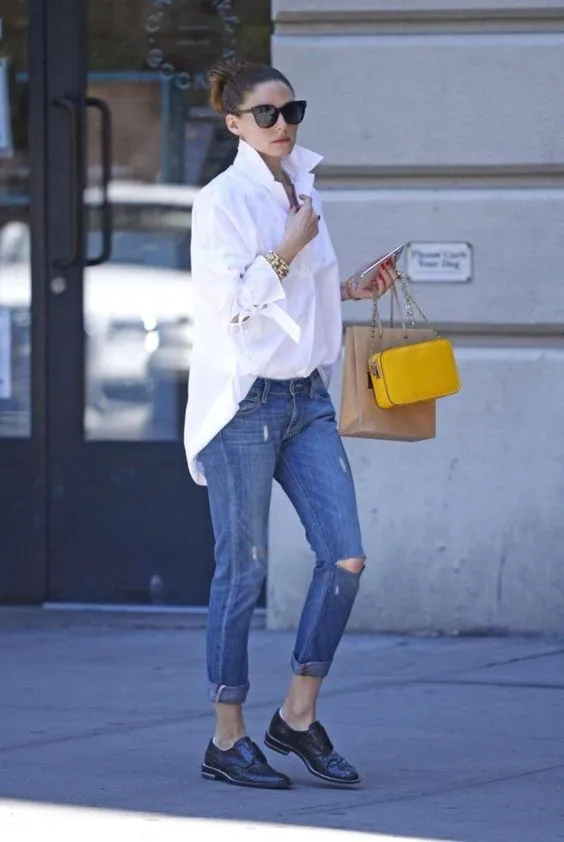 Девушка одета в белую рубашку больших размеров, темно-синие джинсы с потертостями и черные туфли на низком ходу. Образ дополнен аксессуарами, очками и желтой сумкой.