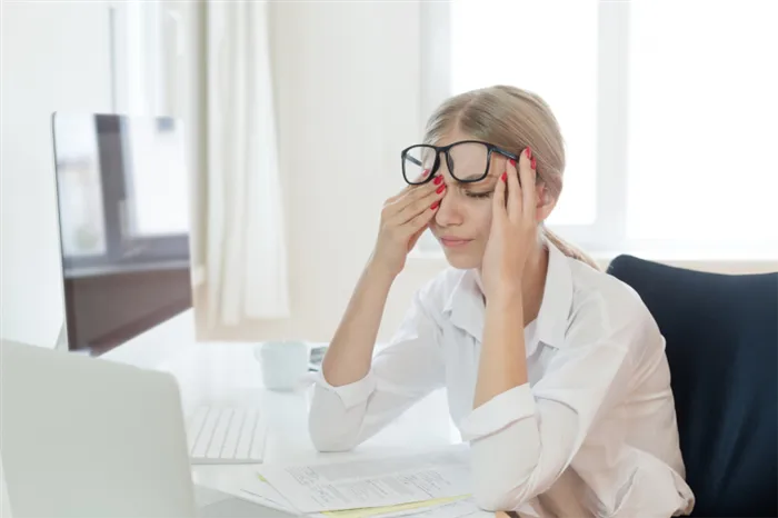 Усталая девушка в белой блузке в офисной обстановке приподнимает очки и трет глаза